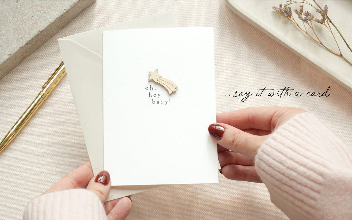 A white greeting card held by two hands, featuring a small wooden shooting star decoration and the text 'oh, hey baby!'. The card is shown with a matching white envelope on a soft beige surface, with a gold pen and dried flowers in the background.